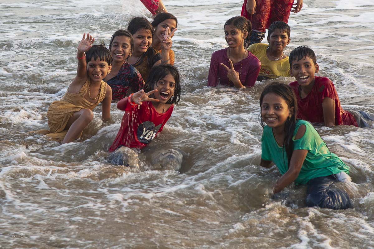 Kinder spielen lachend im Meer, winken in die Kamera und sitzen gemeinsam in den Wellen am Strand.