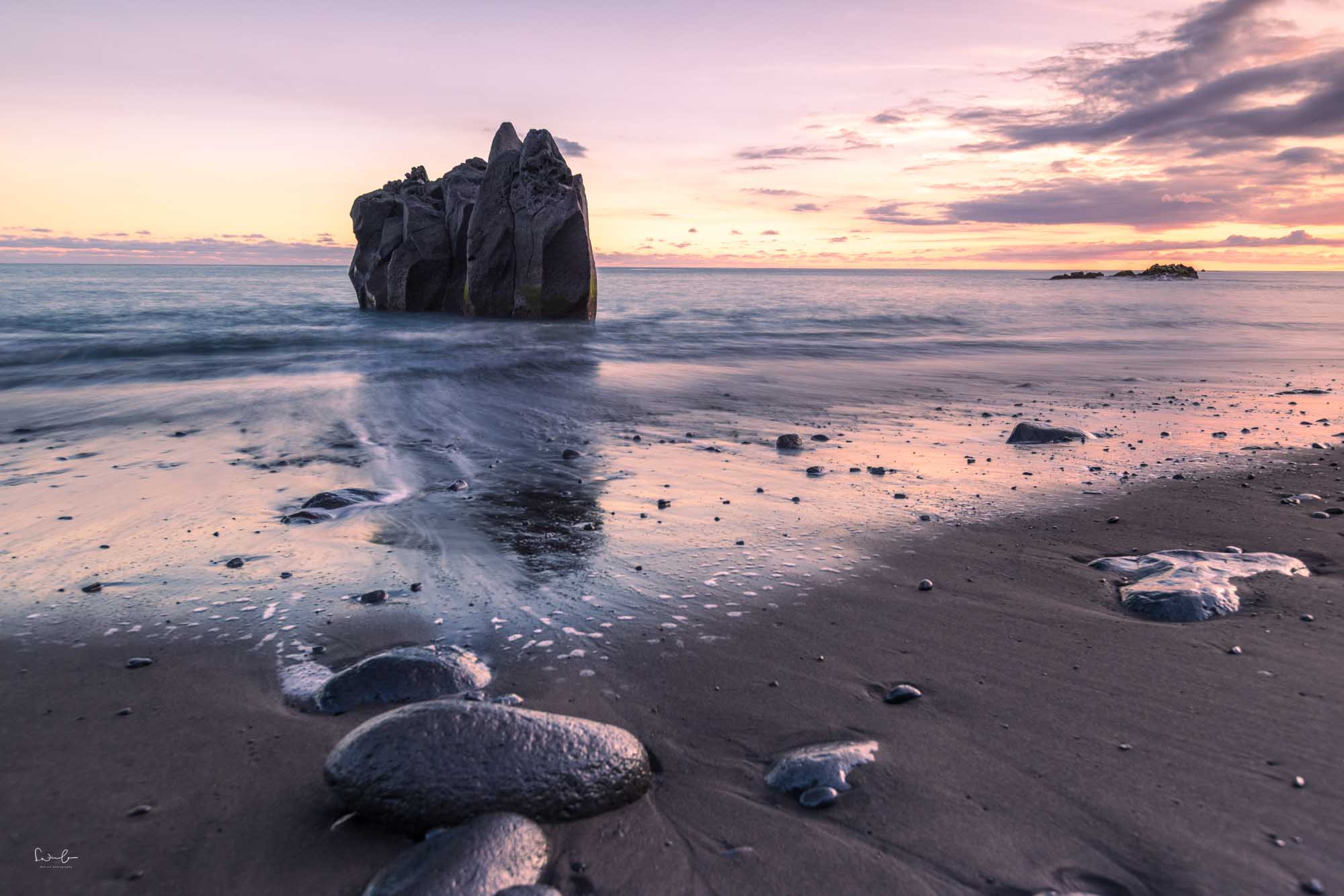 Steine im Vordergrund führen den Blick über einen schwarzen Sandstrand zu einem Felsen im Meer bei Sonnenuntergang.