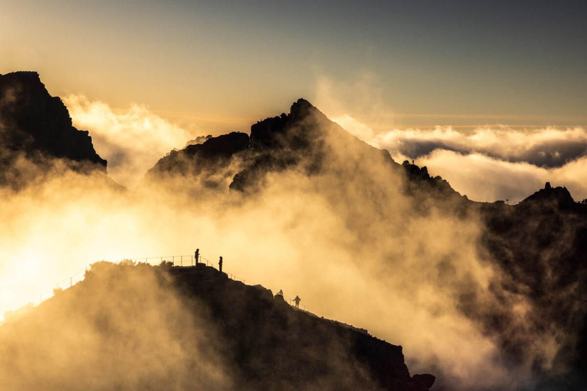 Silhouette von Berggipfeln am Pico do Arieiro auf Madeira bei Sonnenaufgang, goldenes Gegenlicht durchflutet die Wolken zwischen den Felsen und eine kleine Figur steht auf der Aussichtsplattform – ein Beispiel dafür, wie Gegenlicht und die goldene Stunde dramatische Stimmung erzeugen.